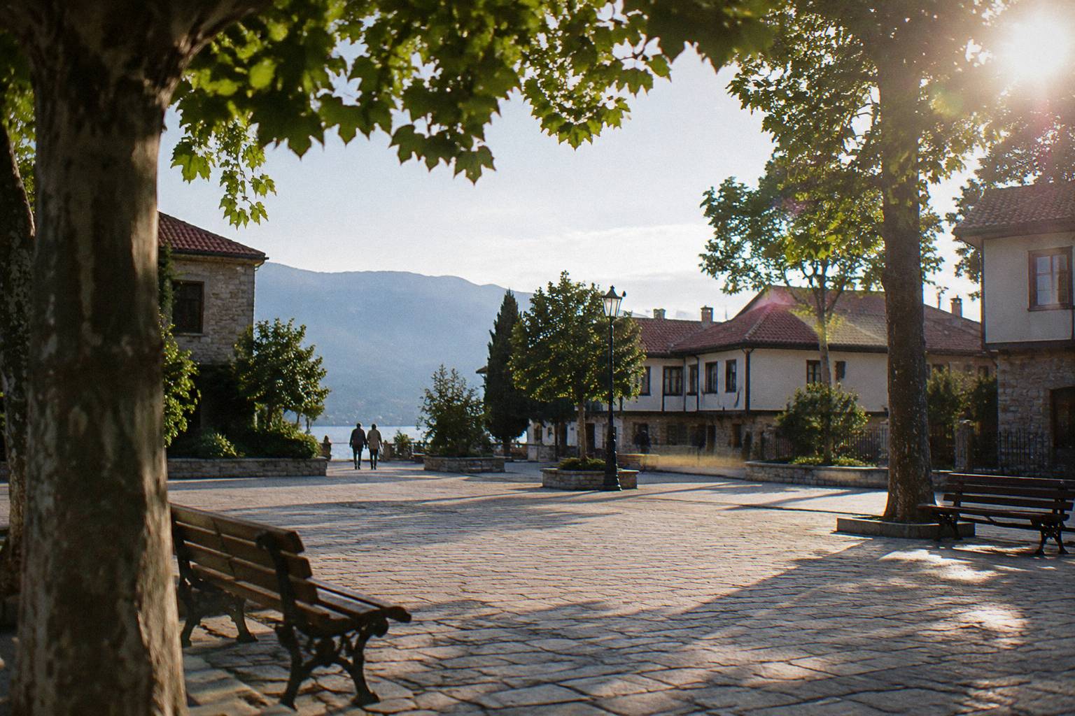 A broad pale-paved square with plane trees, empty cafe-free benches and low old buildings in clear morning light