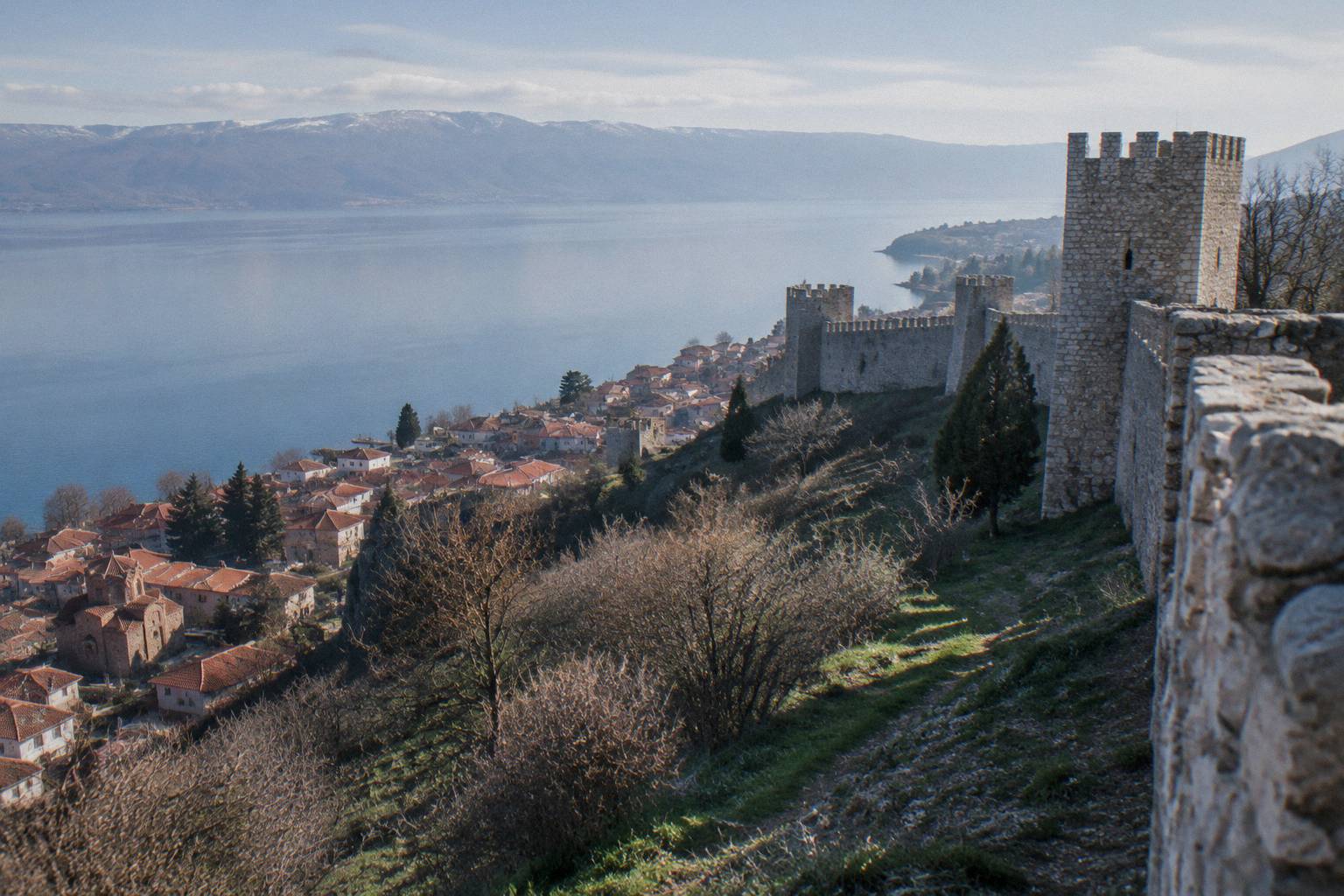 Long weathered stone ramparts and square towers along a ridge above the red roofs of an old lake town in soft morning light