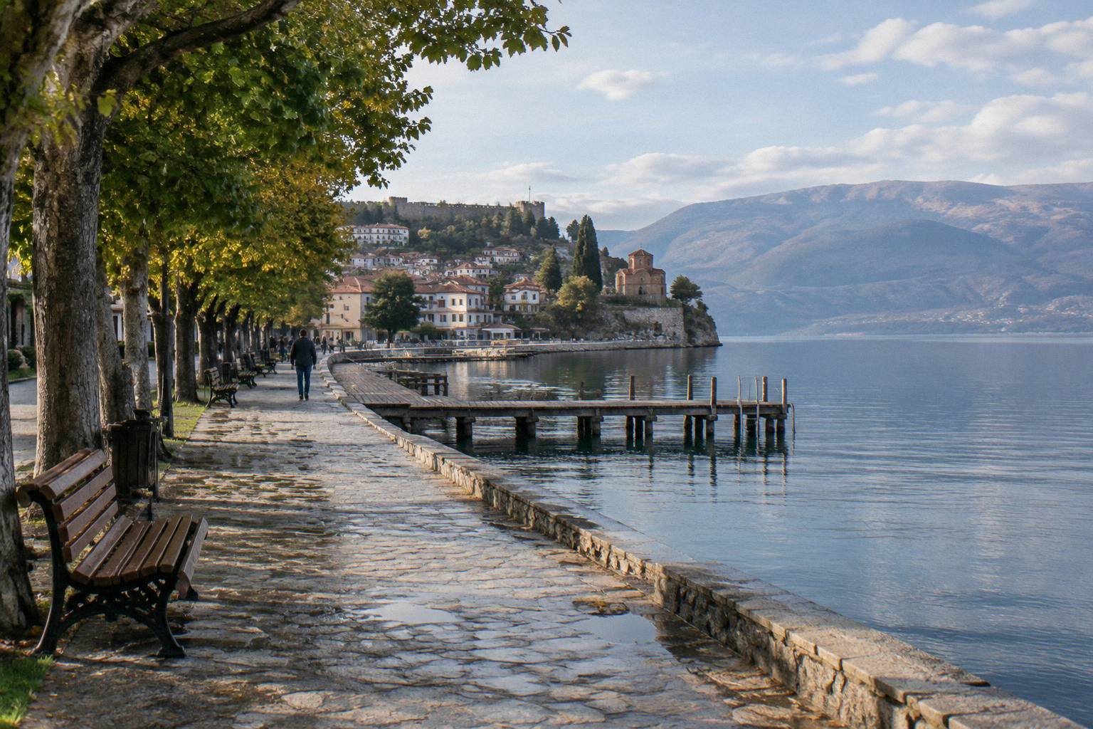 A tree-lined stone lakeside promenade with benches and a wooden jetty on calm water, mountains far across the lake in warm afternoon light