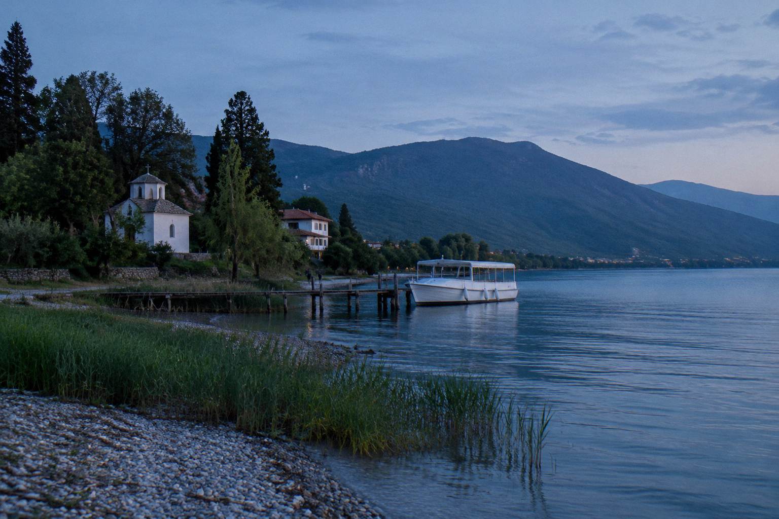 A small white passenger boat moored at a plain wooden jetty on a calm freshwater lake with wooded mountains behind