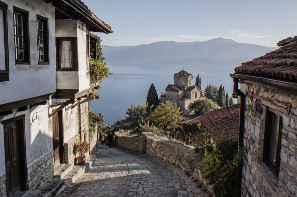 A stone church on a cliff above a calm blue lake with mountains in the distance