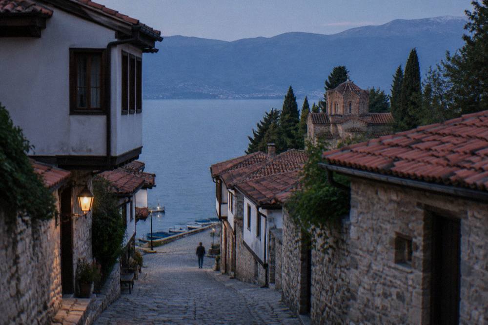 Small boats moored along a lakeside quay with a stone promenade and hills beyond