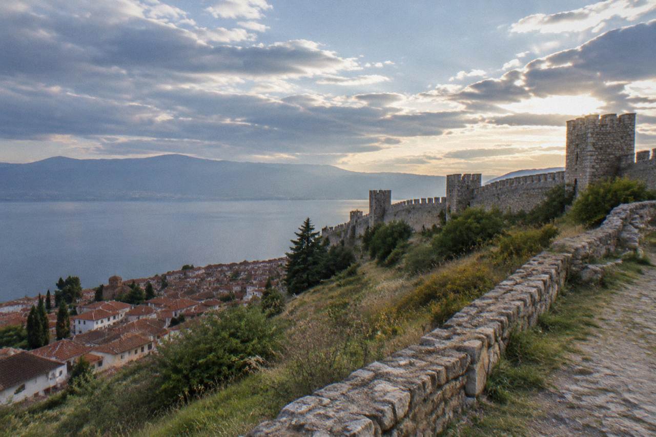 Hillsides above a lakeside old town with red roofs and a stone fortress wall under an evening sky