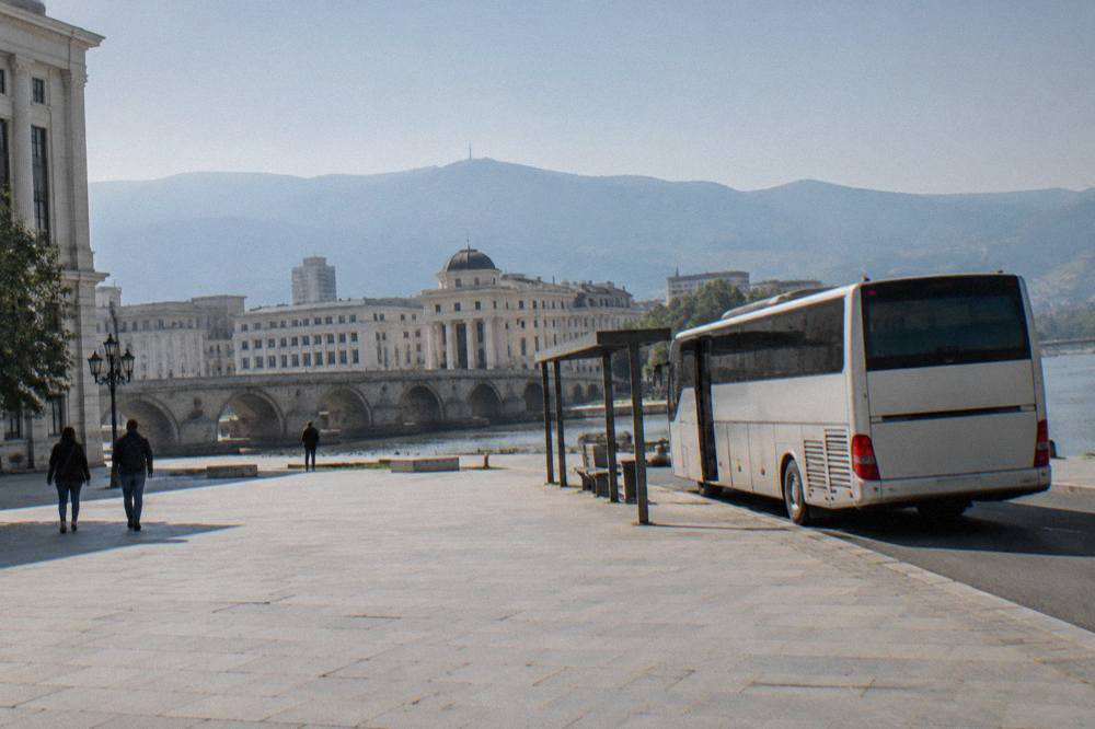 A long-distance bus parked at a small station platform beside a lake road in morning light