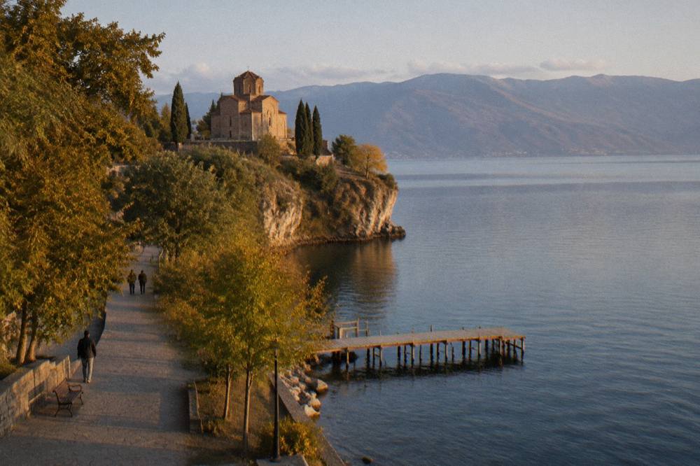 A small church on a cliff above a calm lake with a wooden pier below