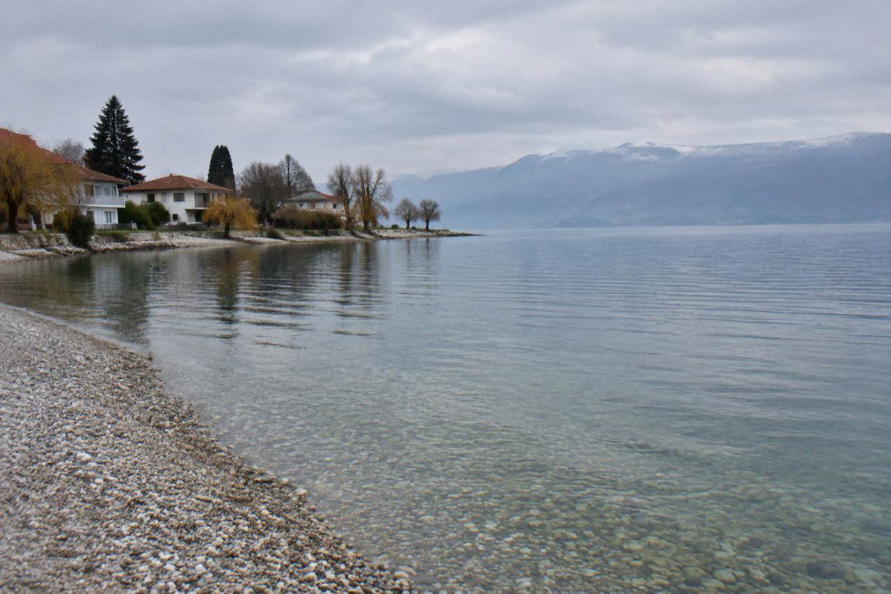 A pebble beach and calm clear water on the Lake Ohrid shore with a few low houses among trees