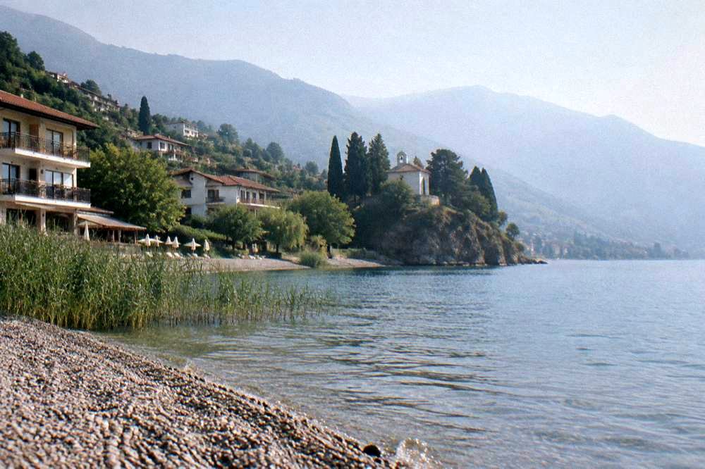 A row of lakeside hotels and guesthouses behind a pebble beach on a bright morning