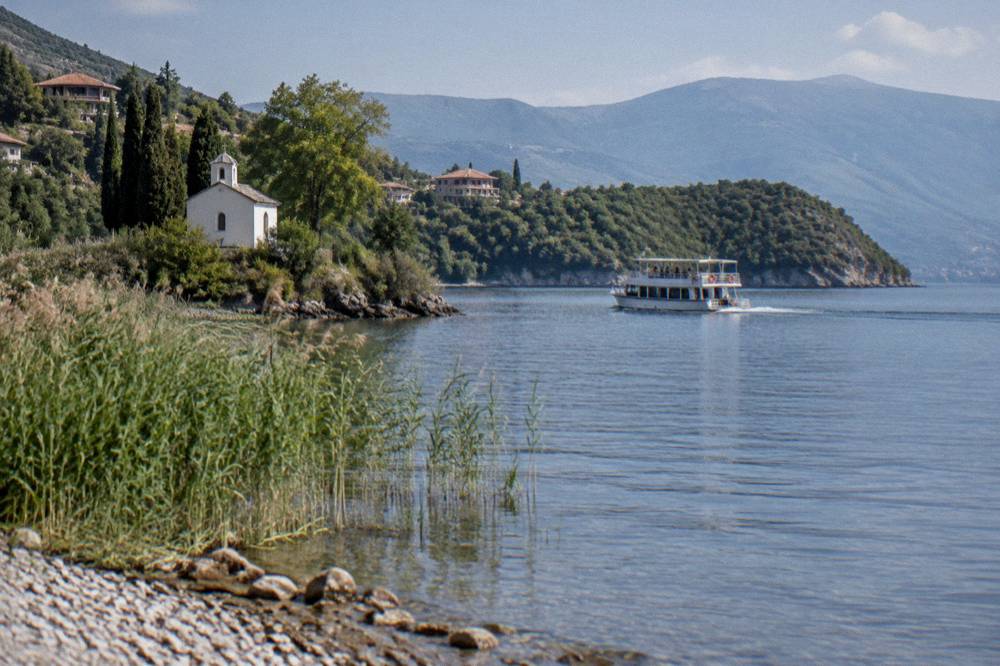 A small cruise boat crossing open water toward a green headland on Lake Ohrid