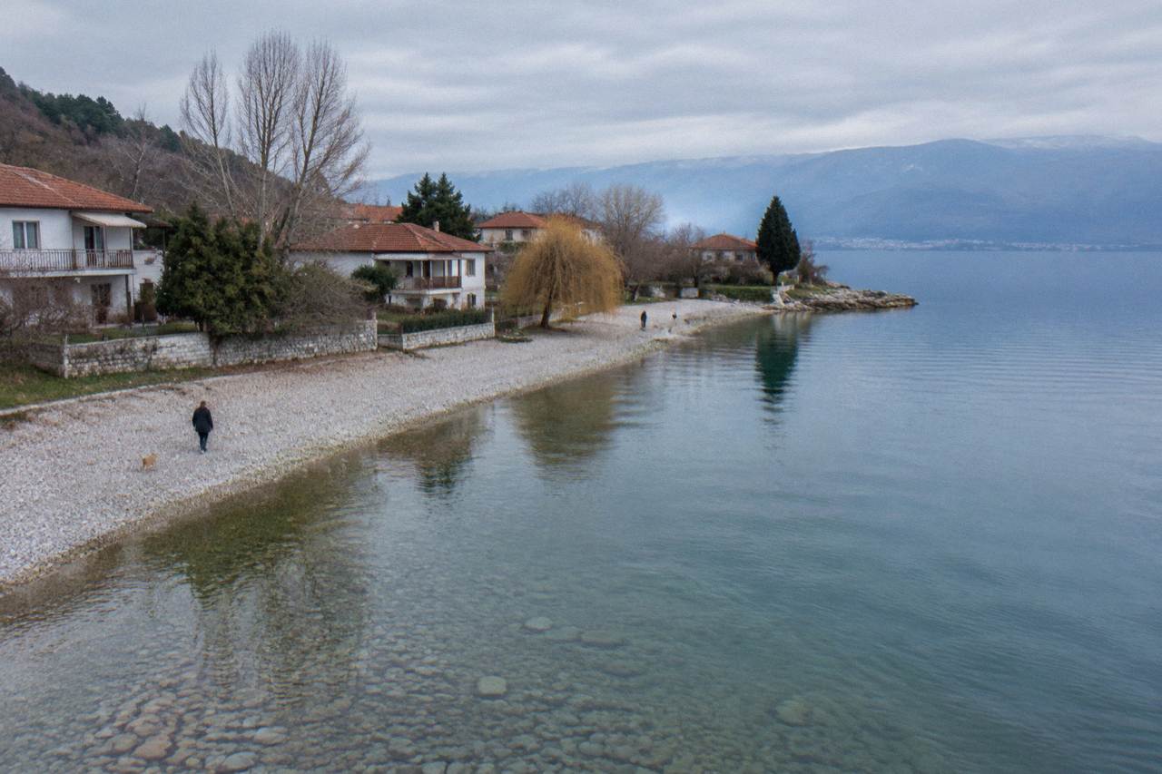 A pebble beach and calm clear water on the Lake Ohrid shore with a few low houses among trees