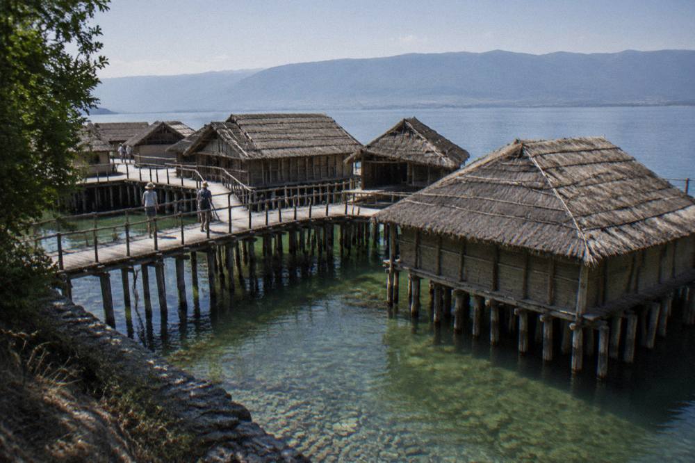 Reconstructed wooden prehistoric lake dwellings standing on pilings above clear green water at the Bay of Bones