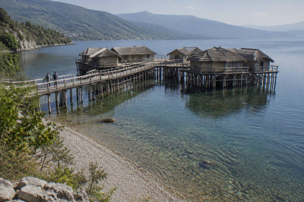 A pebble beach beside clear lake water with wooded hills along the far shore