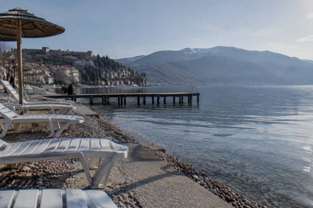 A sunny lake shore with sun loungers and calm water in front of distant mountains