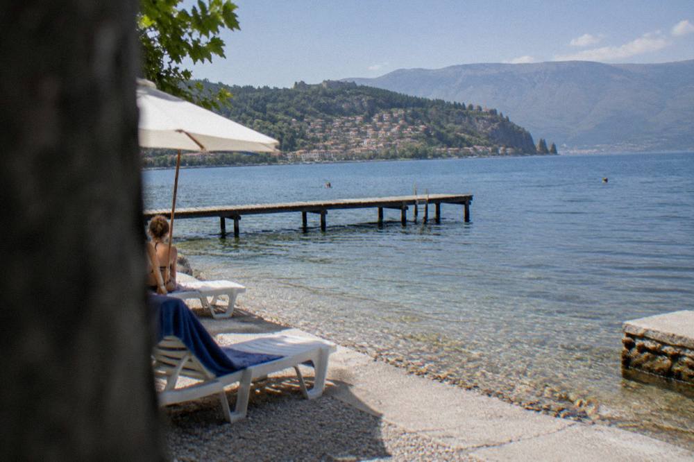 A quiet pebble beach on a lake with wooded hills and a village across the water