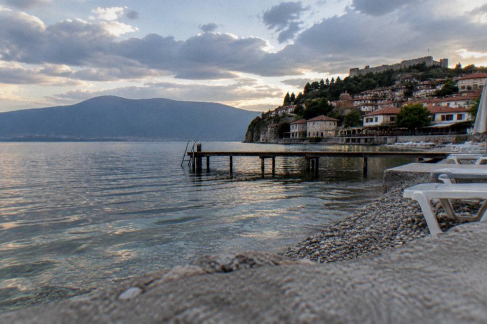 A lakeside town at dusk with lit stone buildings above a calm dark lake