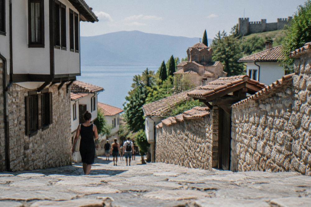 Visitors walking a cobbled street between stone walls below a hilltop fortress in Ohrid