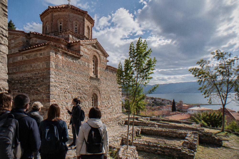 Small tour group listening to a guide beside an old stone church wall in narrow lanes