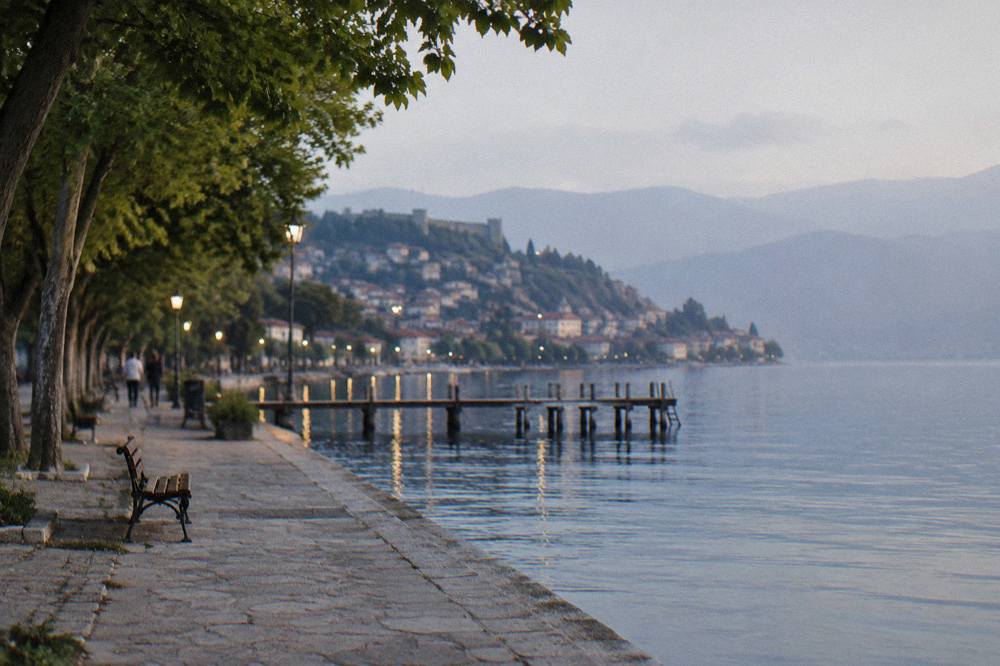 A lakeside promenade at dusk with lights along the water and hills behind