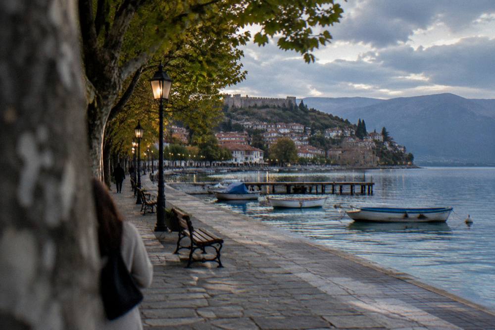 A lakeside promenade at dusk with lanterns lit and moored boats along the shore