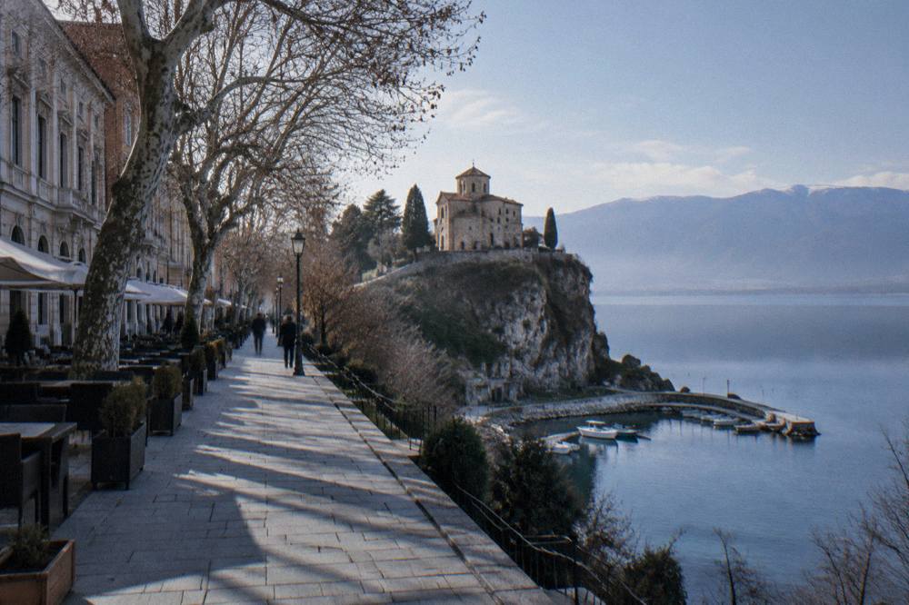 A stone church on a cliff above a calm blue lake with a small harbour below