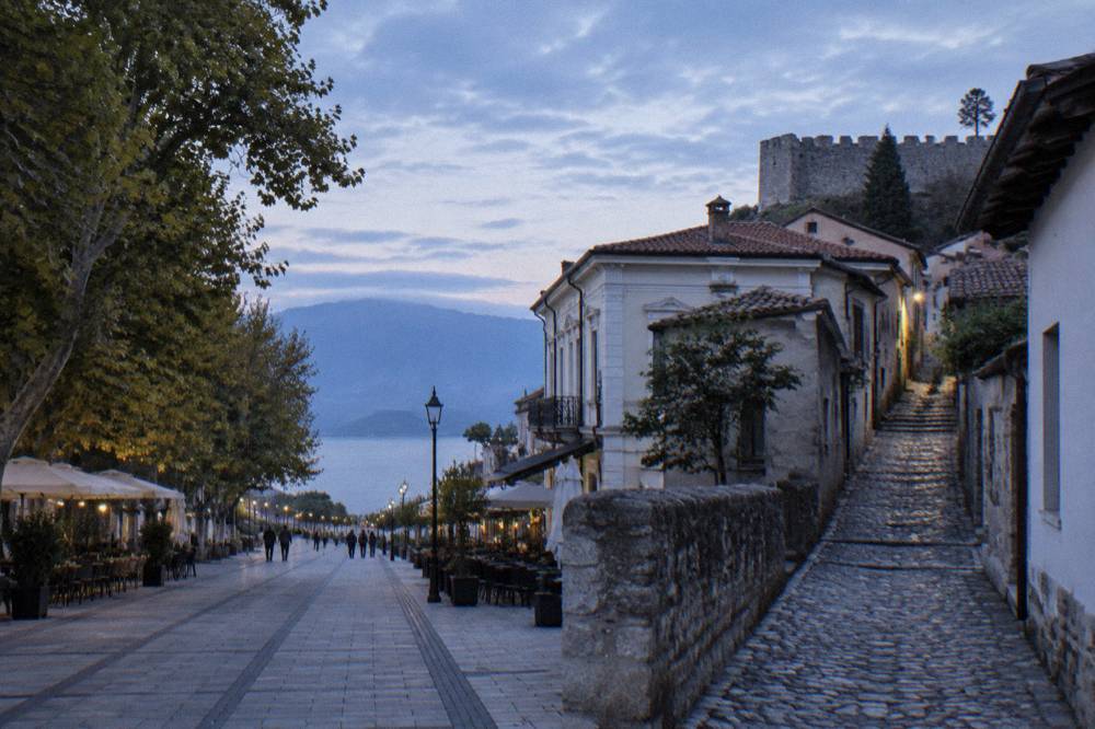 A narrow stone lane climbing between old houses toward a fortress wall above a lake
