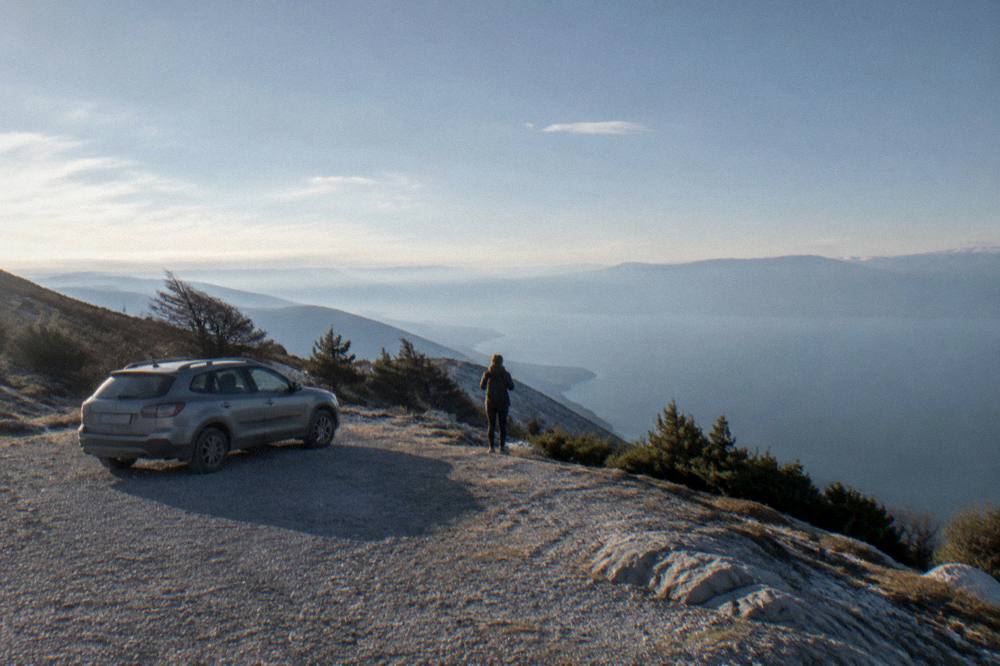 A car parked at a wide gravel pull-off on a mountain road above a hazy lake