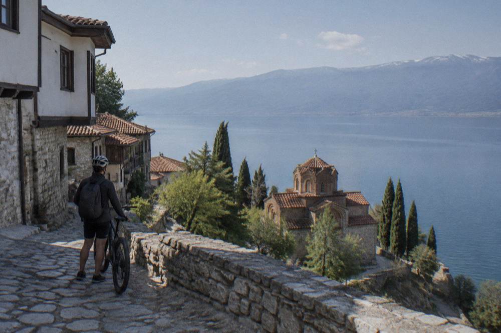 A rider pausing at a lakeside viewpoint above a small church on the shore of Lake Ohrid