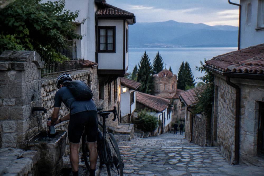 A cyclist filling a water bottle at a fountain on a stone street in Ohrid