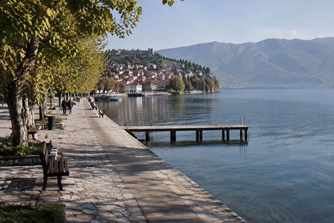 A lakeside promenade on the Albanian shore of Lake Ohrid with a small harbour and hills behind