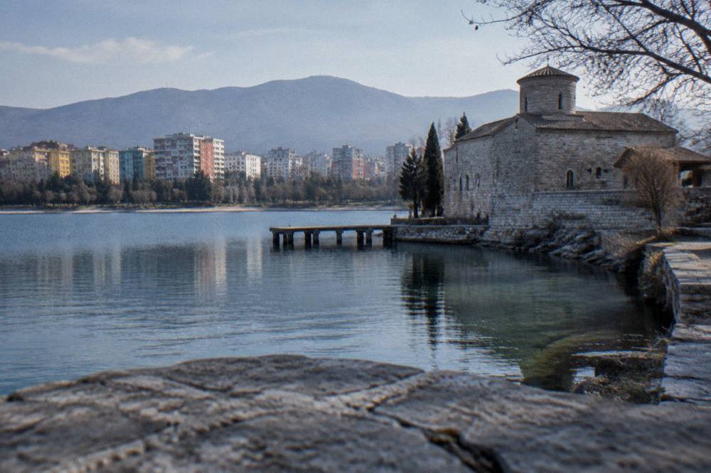 A stone monastery building above a clear lake inlet with a small jetty