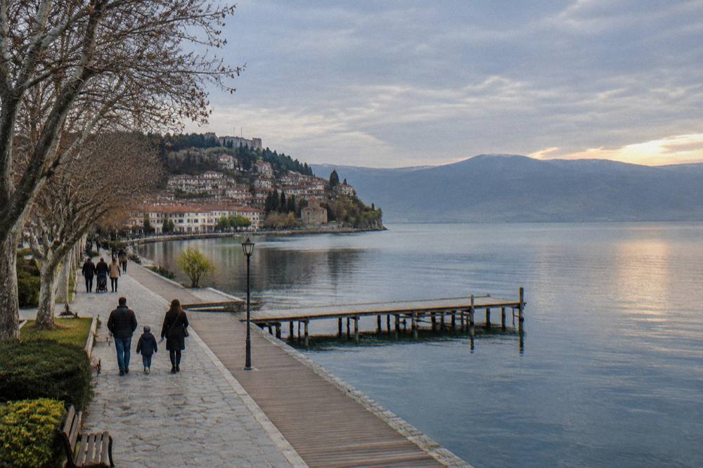 Families walking along a lakeside promenade with trees and calm water at golden hour