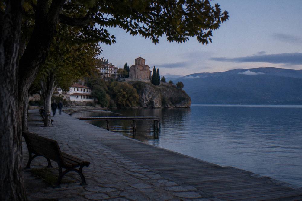 A stone church on a cliff above a blue lake with wooded hills across the water
