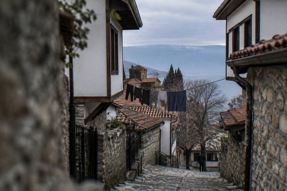 A quiet stone lane in Ohrid's old town with laundry overhead and a distant lake glimpse
