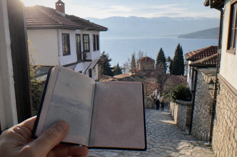 Open passport held in hand at a bright window with a lake town visible in the background