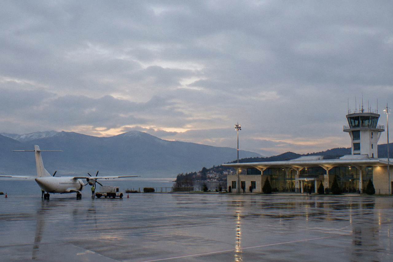 A small regional airport terminal building beside a lake and mountains under an evening sky