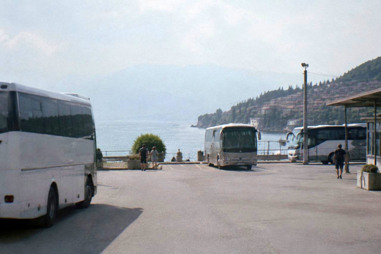 Buses parked at a small station forecourt in a lakeside Balkan town with hills behind