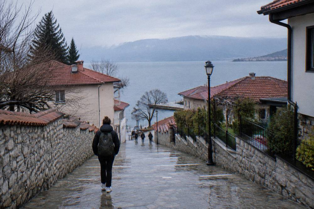 A traveller with a backpack walking down a town street toward a blue lake in the distance