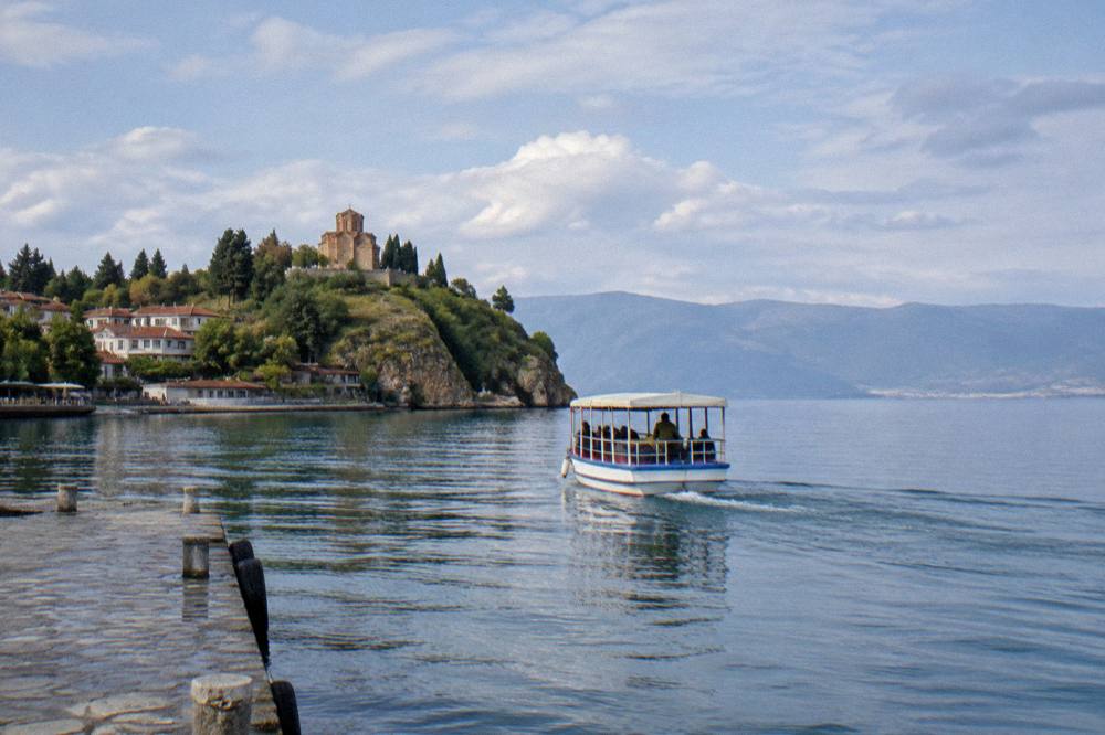 A small passenger boat leaving a lakeside jetty with a hilltop church visible above the shore