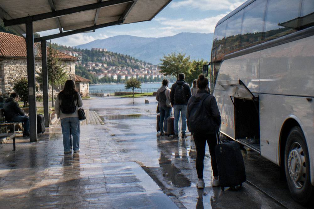 People with bags waiting under a canopy beside a parked coach at a small station