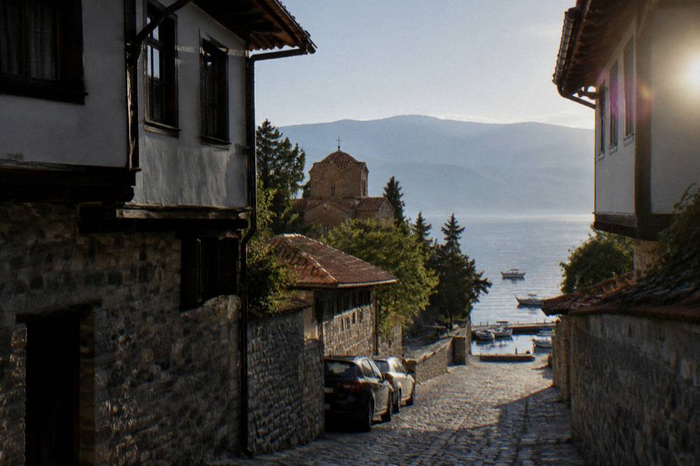 Cars parked in a shaded street near the lake with small boats at a quay beyond
