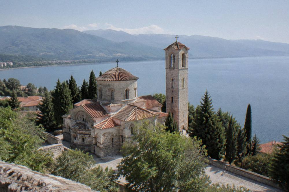 Small stone church with a domed roof and a bell tower among trees above a lake