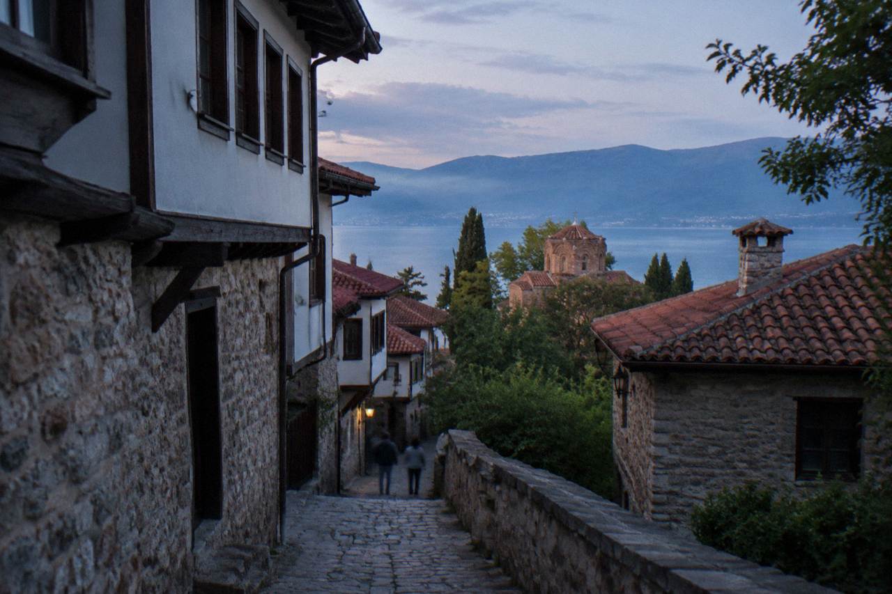 A stone footpath climbing through low trees above the blue water of Lake Ohrid