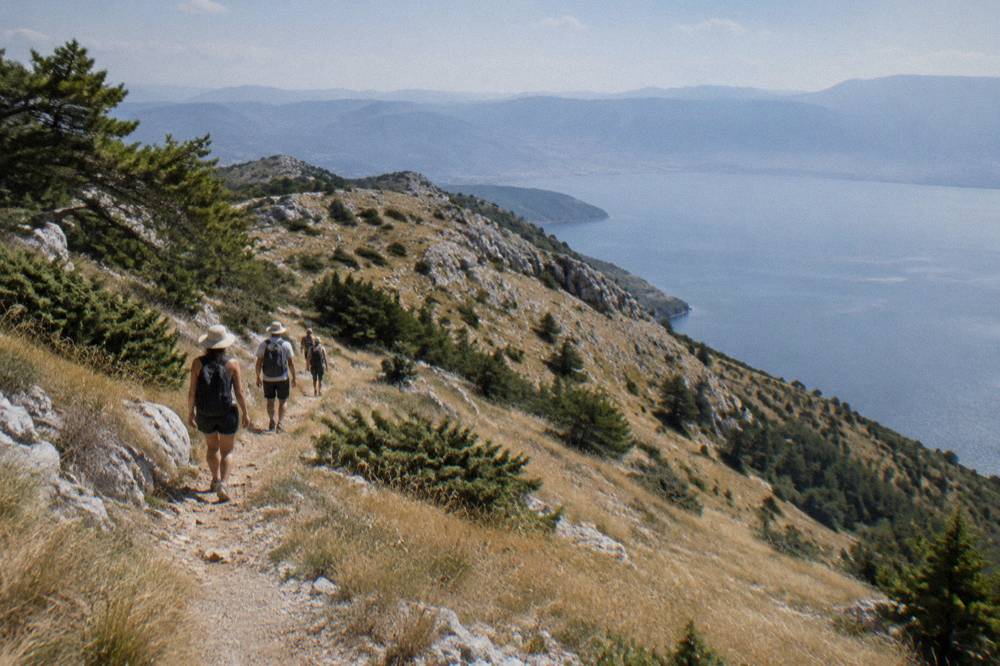 Hikers on a dirt trail through golden summer grass with the lake glittering far below