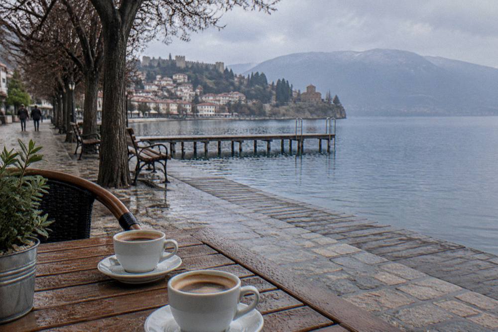 A cafe table with two coffee cups beside a lake promenade in morning light