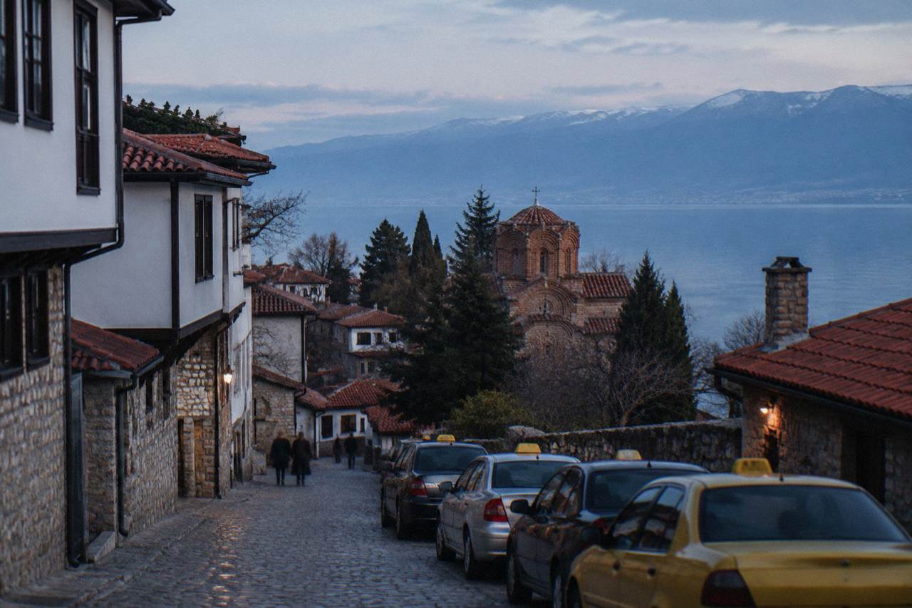 A row of taxis parked on a lakeside street in Ohrid with the old town rising behind them
