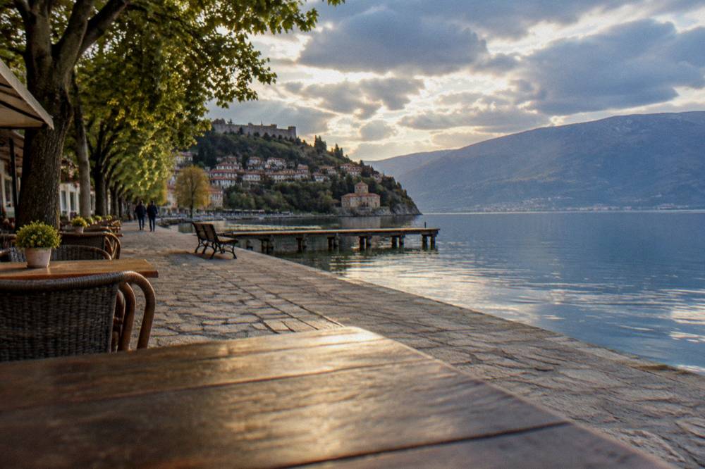 A lakeside promenade in warm evening light with cafe tables and still water reflecting the sky