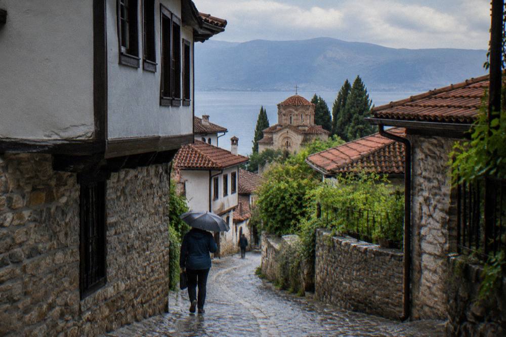 A walker in a rain jacket on a cobblestone lane in Ohrid old town under an umbrella