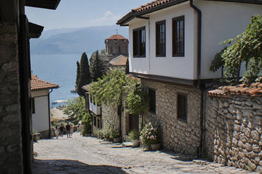 A pebble beach on Lake Ohrid with sun loungers under trees and clear shallow water