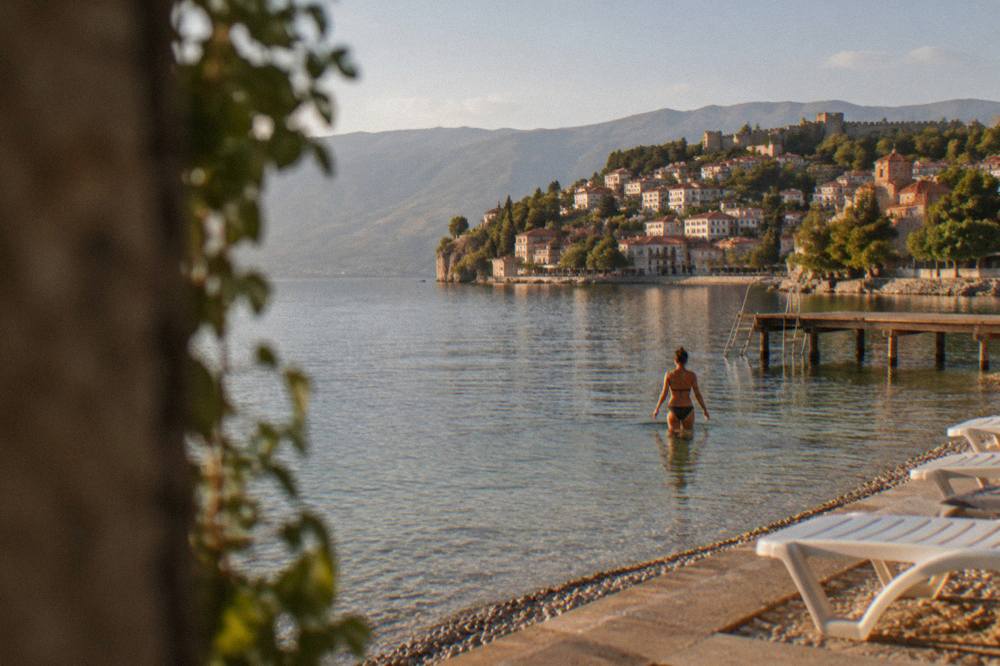 A swimmer walking out of clear lake water toward a stone town on a hill