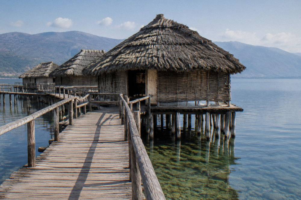 A wooden walkway over lake water leading to a reconstructed prehistoric pile dwelling