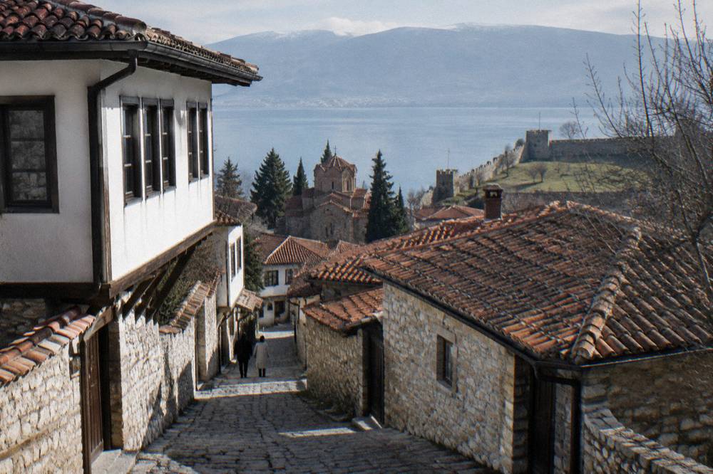 Stone fortification walls on a grassy hilltop above red tiled roofs and a lake